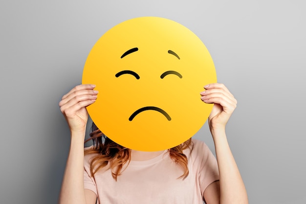 eine blonde Frau hält sich ein trauriges Emoji vor ihr Gesicht